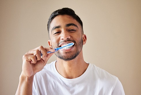 Man with brown hair and white t-shirt brushing teeth with a blue and white toothbrush